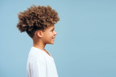 Yan görünüş, gülümseyen, kıvırcık saçlı mutlu Afro-Amerikan çocuk portresi, başka tarafa bakıyor, mavi arka planda izole edilmiş poz, kopyalama alanı. Reklam kavramı