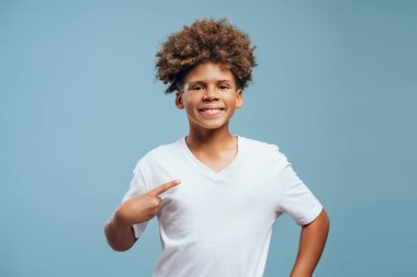 Gülümseyen, neşeli, kıvırcık saçlı, beyaz tişört giyen, kendini gösteren, model olan, kameraya bakan, mavi arka planda yalnız poz veren Afro-Amerikan çocuk. Reklam kavramı