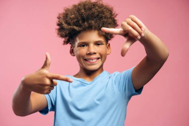Gülümseyen, çekici Afro-Amerikan çocuk portresi, kıvırcık saçlı genç parmaklarından çerçeve yapıyor, pembe arka planda izole edilmiş poz veren kameraya bakıyor. Reklam kavramı