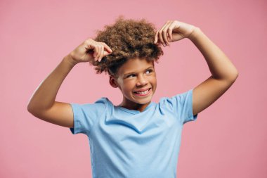 Gülümseyen, neşeli, kıvırcık saçlı, saçlarına dokunan, pembe arka planda soyutlanmış Amerikalı çocuğun portresi. Saç bakım konsepti