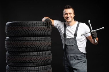 Gülümseyen tamirci İngiliz anahtarı tutuyor ve siyah arka planda bir yığın yeni kış lastiğine yaslanıyor
