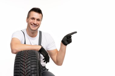 Araba lastiğine yaslanırken ve beyaz arka planda gülümserken parmağıyla işaret eden eldivenler giyen tamirci.