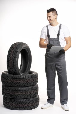 Gülümseyen tamirci, lastik yığınının yanında dururken dijital tablet kullanıyor, envanteri yönetiyor veya oto tamirhanesinde siparişleri kontrol ediyor