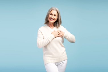 Güzel, mutlu, yaşlı, gri saçlı bir kadının portresi. Günlük giysiler giymiş, kalbe el ele tutuşmuş, kameraya bakıyor, mavi arka planda izole bir şekilde poz veriyor. Minnettar konsept