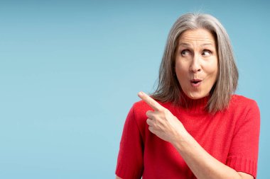 Aşırı mutlu, gülümseyen olgun bir kadının portresi kırmızı süveter giyen, fotokopi alanını işaret eden, mavi arka planda izole edilmiş. Reklam kavramı