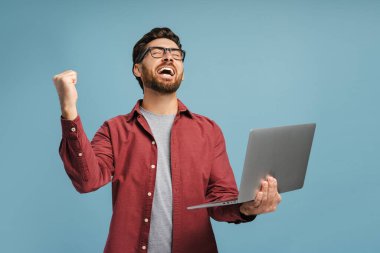 Gözlük takan, dizüstü bilgisayar kullanan, para kazanan, mavi arka planda izole edilmiş spor bahisleri oynayan mutlu, sakallı bir adamın portresi. Çevrimiçi teknoloji kavramı