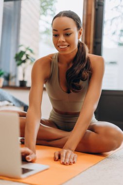 Laptopun önünde yoga yapan gülümseyen Afrikalı Amerikalı portresi, evdeki spor minderinin üzerinde oturuyor. Sağlıklı yaşam tarzı, spor anlayışı