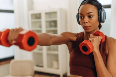 Sportif Afrikalı Amerikalı kadın portresi kulaklıkla müzik dinliyor evde halterlerle egzersiz yapıyor, boks yapıyor, kol kaslarını güçlendiriyor.