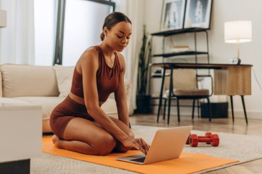 Spor minderinde yoga yapan, evdeki oturma odasında dizüstü bilgisayar kullanan güzel Afrikalı Amerikalı kadın portresi. Sağlık, sağlıklı yaşam tarzı konsepti