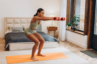 Genç, güzel, Afro-Amerikan bir kadın, dambılları yatak odasında eğitip, çömeliyor. Spor, sağlık hizmeti kavramı