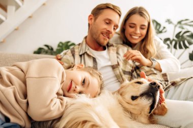 Gülümseyen güzel bir aile, genç bir anne, baba ve küçük bir kız birlikte rahatlıyor, kadın kucaklayan bir adam, küçük kız okşuyor ve köpekle oynuyor, golden retriever. Aşk kavramı, ebeveynlik.