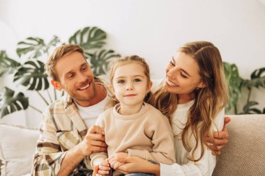 Mutlu, güzel aile portresi, küçük kız, anne ve baba kucaklaşıyorlar evdeki oturma odasında rahat bir kanepede oturuyorlar. Ebeveynlik, çocukluk kavramı