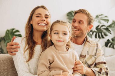 Güzel, gülümseyen aile portresi, küçük kız, anne ve baba kucaklaşıyorlar evdeki oturma odasında rahat bir kanepede oturuyorlar. Aşk, çocukluk kavramı