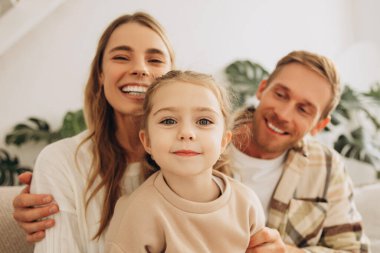 Gülümseyen, mutlu bir aile, güzel bir anne, çekici bir baba ve küçük bir kızın kucaklaşması, oturma odasında rahat bir koltukta oturup kameraya bakması. Çocukluk, ebeveynlik kavramı