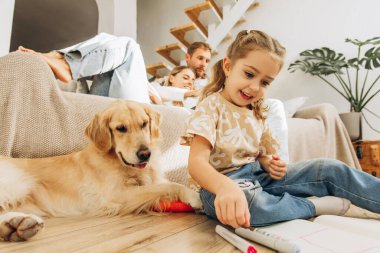 Mutlu bir aile, güzel bir kadının kocasına sarılması, küçük bir kızın çizimi, köpekle yerde oturması, Golden Retriever, rahat bir ev. Uyum kavramı, ebeveynlik, özen.