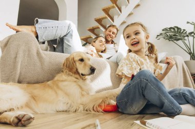 Gülümseyen küçük bir kızın portresi. Oturma odasında yerde oturan köpek, golden retriever ve mutlu ebeveynlerin kucaklaşması. Hayvan sevgisi konsepti