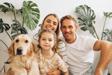 Güzel bir ailenin portresi, mutlu bir anne, baba, sevimli, küçük bir kızın köpekle kucaklaşması, Golden Retriever 'ın evinde, rahat bir oturma odasında. Hayvan bakımı konsepti
