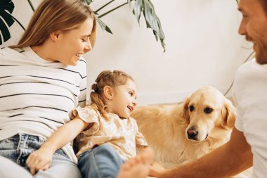 Güzel, mutlu bir aile, çekici bir kadın, köpekle oynayan bir adam ve küçük bir kız, evdeki rahat oturma odasında Golden Retriever. Ebeveynlik, hayvan bakımı konsepti