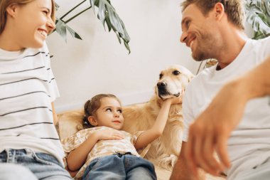 Mutlu bir ailenin portresi, çekici bir kadın, köpekle oynayan bir adam ve küçük bir kız, evdeki sıcak oturma odasında Golden retriever. Ebeveynlik, hayvan bakımı konsepti