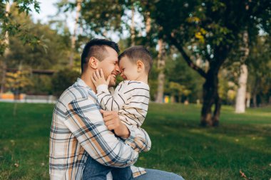Gülümseme portresi, mutlu baba ve küçük oğlun kucaklaşması, parktaki yeşil çimlerin üzerinde battaniyenin üzerinde oturması. İlişki, çocukluk kavramı