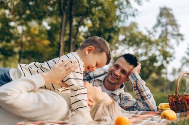 Parktaki yeşil çimlerin üzerinde battaniyeye oturup piknik yapan güzel anne, baba ve küçük oğlunun portresi. Ebeveynlik, çocukluk kavramı