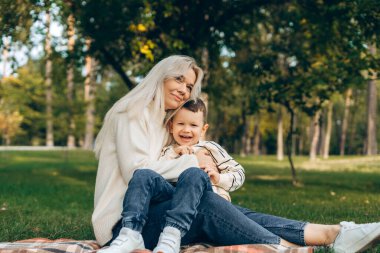 Gülümseme portresi, güzel anne ve küçük oğlu birlikte battaniyeye oturmuş, parktaki yeşil çimlerde dinleniyorlar. Annelik kavramı
