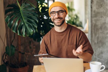 Mutlu, yakışıklı, orta yaşlı, şık sarı şapkalı dijital tablet giyen, kameraya bakan, el kol hareketi yapan, dizüstü bilgisayar kullanan bir adamın portresi. Çevrimiçi teknoloji kavramı