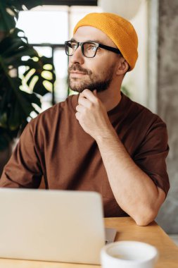 Başarılı, düşünceli, yakışıklı sakallı bir adamın portresi, modern çalışmalarda dizüstü bilgisayar kullanarak şık sarı şapkalı ve gözlüklü bir hippi. İş konsepti