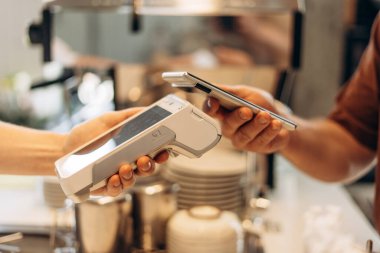 Customer is holding his smartphone over a payment terminal, making a contactless payment for his coffee at the counter of a coffee shop clipart