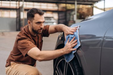Ciddi sakallı, yakışıklı bir adamın portresi. Benzin istasyonunda beyaz SUV 'yi mikrofiber bezle siliyor. Araba bakımı konsepti