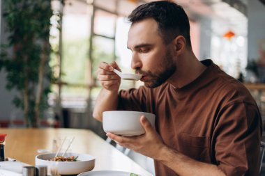 Düşünceli sakallı bir adamın portresi, Çin yemeği tabağı ve kaşığı yiyor, modern kafede oturuyor, öğle yemeği yiyor. Yiyecek konsepti