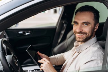 Mutlu, gülümseyen, başarılı iş adamı dijital tablet tutuyor, online çalışıyor, modern yeni arabada oturuyor, kameraya bakıyor. Ulaşım kavramı