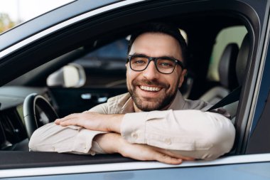 Mutlu, gülümseyen, şık gözlükler takan, modern bir arabada oturan, kameraya bakan Latin bir adam. Araç, sigorta kavramı