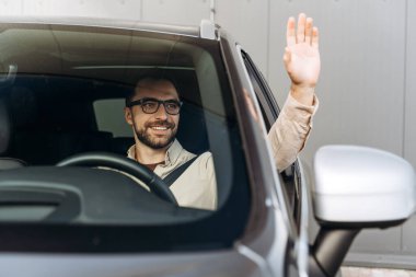 Kendine güvenen, gülümseyen, şık gözlükler takan modern araba kullanan, el sallayan ve direksiyonu tutan İspanyol bir adamın portresi. Sigorta konsepti