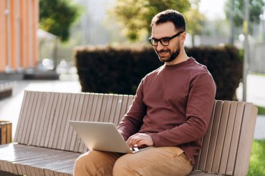 Gülümseyen, sakallı, gözlük takan, dizüstü bilgisayar kullanan, sokakta online alışveriş yapan bir adamın portresi. Çevrimiçi teknoloji kavramı