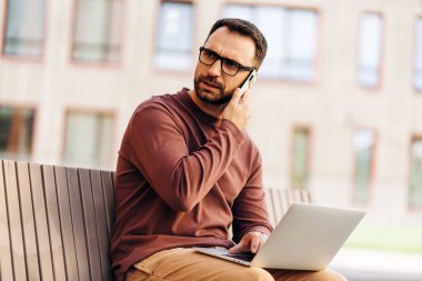 Ciddi, düşünceli, gözlük takan, laptop kullanan, telefonlara cevap veren, dışarıda bankta oturan sakallı bir adamın portresi. Çevrimiçi sosyal medya kavramı