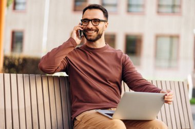 Gülümseyen, gözlüklü, gözlüklü yakışıklı bir adamın portresi, parkta bankta oturuyor, dizüstü bilgisayar kullanıyor, telefona cevap veriyor, cep telefonu tutuyor. Çevrimiçi iletişim kavramı