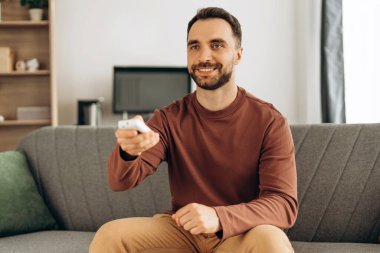 Uzaktan kumandalı, mutlu, sakallı, sakallı bir adamın portresi. Video, televizyon izliyor, evdeki rahat kanepede oturuyor. Hobi konsepti