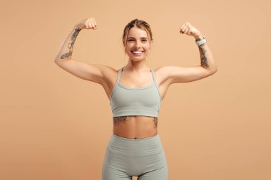Spor elbiseli gülümseyen genç bir kadının portresi kollarındaki kasları gösteriyor, stüdyoda bej arka planda poz veriyor. Spor, sağlık hizmeti kavramı