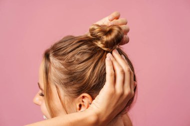 Saçını topuz yapan, stüdyoda poz veren, pembe üzerine izole edilmiş gülümseyen genç bir kadın. Moda ve kuaför konsepti