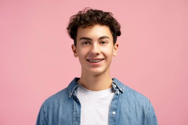 Diş teli takan, günlük gömleği olan mutlu bir çocuk pembe arka planda izole edilmiş kameraya bakıyor. Reklam kavramı