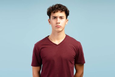 Ciddi, düşünceli, kırmızı tişört giyen, kameraya bakan, mavi arka planda tek başına duran genç bir çocuğun portresi. Olumsuz duygular konsepti