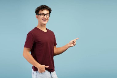 Gözlüklü, mutlu ve yakışıklı bir gencin portresi mavi arka planda tek başına duran fotokopi alanını işaret ediyor. Reklam kavramı