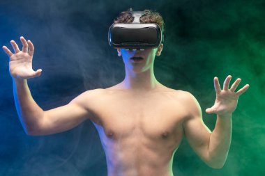 Çıplak gövdeli, VR gözlüklü, online video oyunu oynayan, mavi dumanlı arka planda tek başına duran bir çocuk. Çevrimiçi teknoloji kavramı