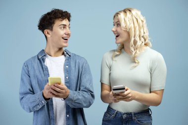 Heyecanlı, gülümseyen arkadaşlar, ergenler, günlük kıyafetler giyen kız ve erkek ellerinde cep telefonları, mobil uygulama kullanıyorlar, birbirlerine bakıyorlar, mavi arka planda tek başlarına duruyorlar. Çevrimiçi teknoloji kavramı