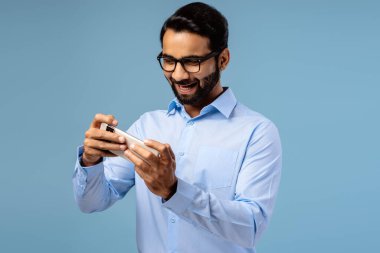 Heyecanlı orta yaşlı gözlüklü Hintli adamın portresi, cep telefonu tutan, video oyunu oynayan, mavi arka planda tek başına duran oyuncu. Çevrimiçi teknoloji kavramı