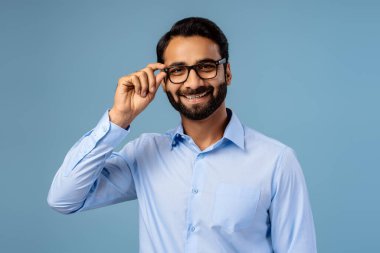 Yakışıklı, gülümseyen, mutlu, sakallı, gözlüklü bir adam mavi arka planda izole edilmiş kameraya bakıyor. Görüş konsepti