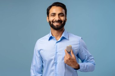 Portrait of sick, smiling Indian man, patient holding pacemaker, looking at camera, posing isolated on blue background. Health care, cardiology concept clipart