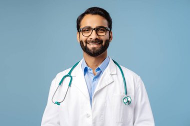 Gülümseyen, çekici Hintli sakallı adam, şık gözlükler takan doktor, kameraya ve beyaz cekete bakan, mavi arka planda tek başına duran kameraya bakan. Sağlık hizmeti kavramı