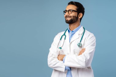 Yakışıklı, güler yüzlü, kendine güvenen Hintli profesyonel doktor beyaz önlüklü ve kolları çaprazlanmış gözlüklü, mavi arka planda izole edilmiş fotokopi alanına bakan. Tıbbi konsept